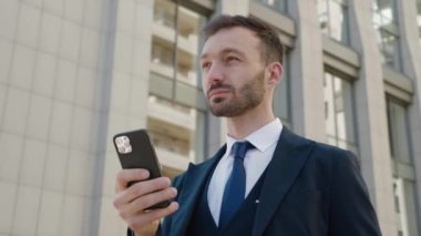 Modern Office binasının yakınındaki şehir merkezinde duran akıllı telefonu kullanan çekici iş adamının portresi. Klasik takım elbise giyiyordu. Sosyal ağ. Uygulamalar. Akıllı telefonlar.