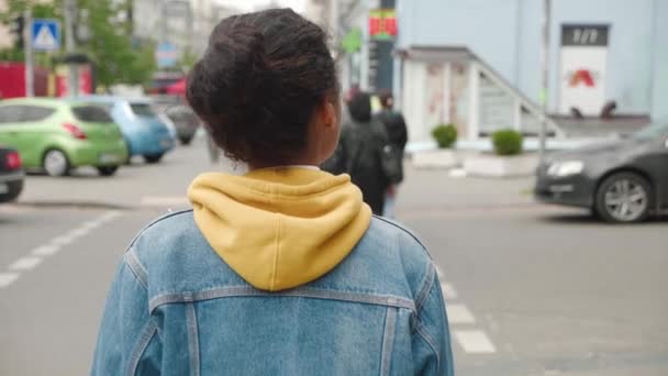 Vista trasera de la elegante mujer afroamericana en una chaqueta de jean caminando ciudad en el centro de la ciudad. Vista trasera de la joven estudiante que cruza la calle con gente borrosa en el fondo, la vida ocupada de la ciudad. — Vídeo de stock