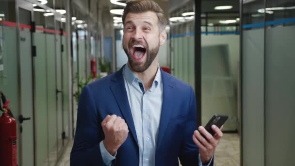 Excited young businessman rejoices at message on his smartphone, standing at office. Happy male looking at mobile phone celebrate good news victory success. Lucky man showing yes winner gesture. — стоковое видео