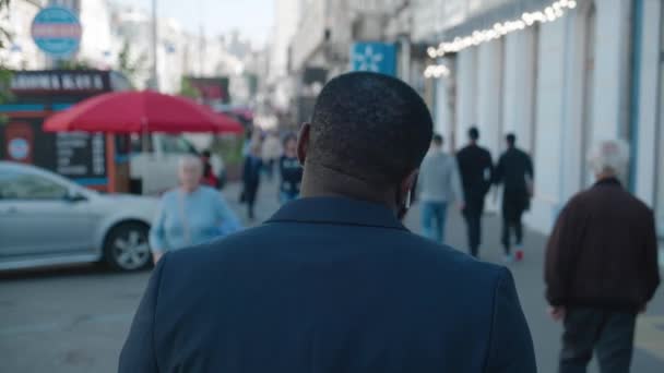 Back view of unrecognizable african american man walks along the city center street looks around city on background business downtown. Rear back view of businessman wearing suit, commuting to work. — Stok Video