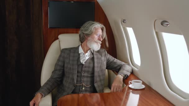 Homme millionnaire mature élégant portant une combinaison classique de luxe volant dans un avion privé. Homme d'affaires PDG confiant regardant par la fenêtre du jet d'entreprise. Voyager de luxe en jet privé. — Video