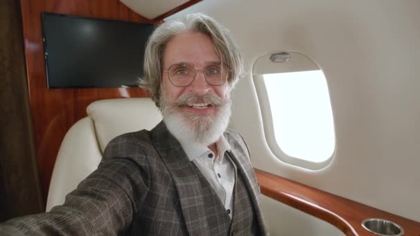 Happy mature millionaire man talking to camera holding phone recording vlog while flying on private jet. Gray-haired stylish senior man video calling in online chat using smartphone at airplane. — Stock Video