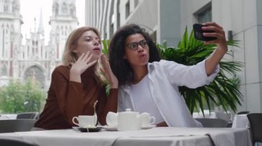 İki farklı kadın arkadaş cep telefonlarıyla selfie çekiyorlar, gülümsüyorlar ve şık bir sokak kafesinde otururken eğleniyorlar..
