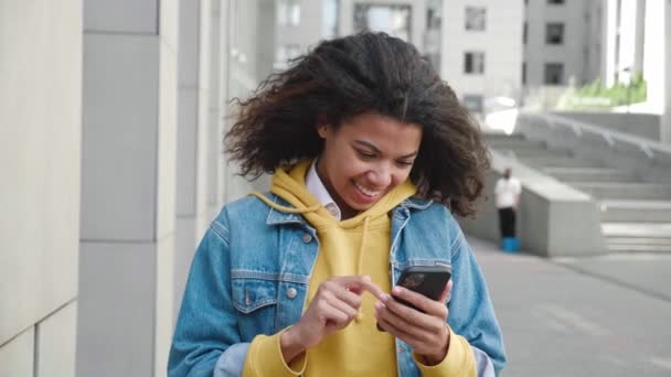 Retrato de mujer adolescente de raza mixta sonriente usando la aplicación en el teléfono inteligente, de pie al aire libre en la calle de la ciudad — Vídeo de stock
