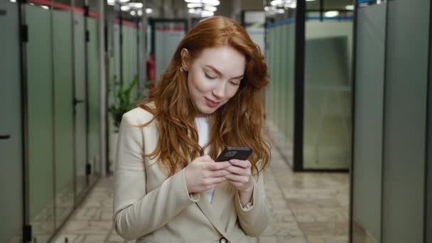 Sourire attrayant message texte femme d'affaires sur son smartphone. Portrait de femme exécutive confiante avec les cheveux roux dans l'usure formelle en utilisant l'application sur téléphone mobile tout en se tenant dans le couloir de bureau. — Video