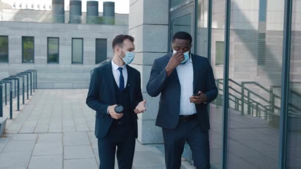 Casal de dois empresários diversos conversando e andando fora do prédio de escritórios durante o período de quarentena. Dois funcionários colegas de trabalho trabalhadores gerente em ternos usando máscaras protetoras durante o confinamento — Vídeo de Stock