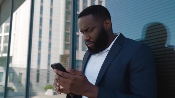 Jistý vousatý afro-americký muž procházení internetu na moderním smartphonu, zatímco stojí v blízkosti kancelářského centra. Portrét obchodníka, který si dává pauzu od práce venku s kávou v centru — Stock video
