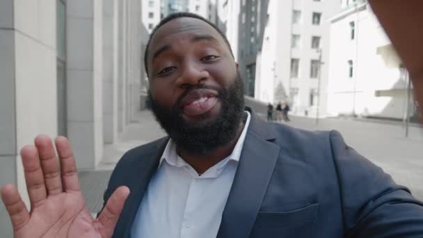 Sonriente hombre de negocios afroamericano haciendo videollamada selfie de pie al aire libre en el distrito de negocios. Retrato de un bloguero de negocios filmando video blog al aire libre cerca del moderno edificio de oficinas. — Vídeos de Stock