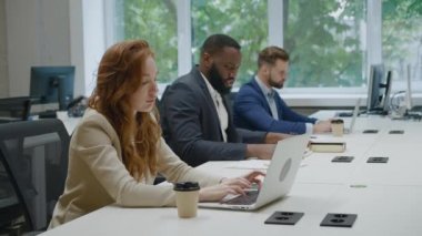 Modern ofisteki bilgisayarda çalışan genç ve çekici kızıl saçlı iş kadınının portresi. Ofis çalışanları arka planda.
