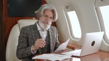 Özel bir jet uçağında uçarken dokümanlarla uğraşan ve dizüstü bilgisayar kullanan yetişkin bir işadamı..