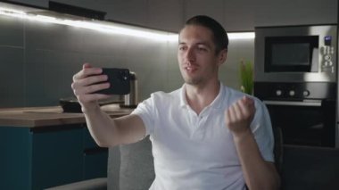 Gülümseyen genç adam evdeki kanepede otururken video görüşmesi yapıyor. Erkek portresi yaşam arka planında görüntülü konuşma yapıyor.