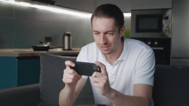 Akıllı telefonuyla oynayan genç adam heyecan verici mobil uygulaması evdeki oturma odasında kanepede otururken yumruğunu sıkarak bir yarış oyunu kazanıyor..