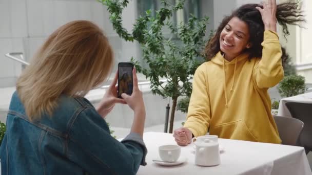 En kvinna fotograferar en annan kvinna sin vän på smartphone kamera i mysiga utomhus café. Positivt leende ung blandad ras elegant flicka poserar för foto på gatan café — Stockvideo