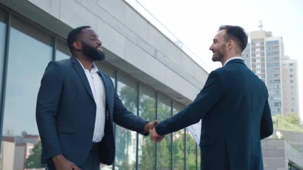 Dos hombres de negocios se dan la mano cuando se reúnen, aceptan un trato o saludan. Retrato de apretón de manos de dos hombres de negocios sonrientes haciendo un trato. Edificio de oficinas en segundo plano. Distrito de negocios. — Vídeo de stock