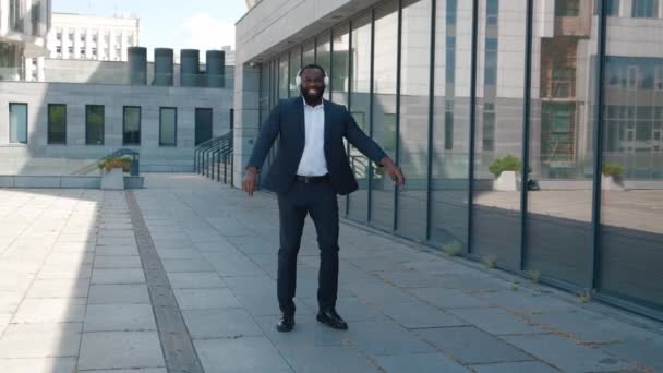 Позитивный африканский американский бизнесмен в официальном костюме с наушниками, танцующими и поющими на открытом воздухе — стоковое видео