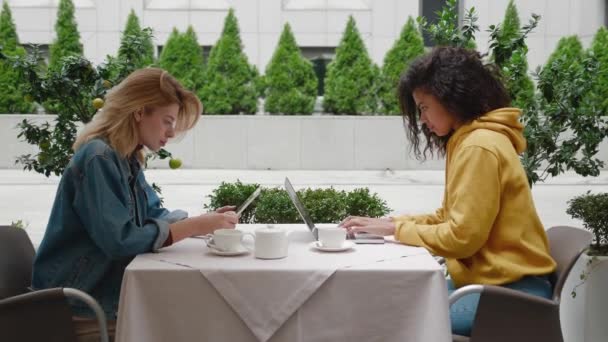 Zwei unterschiedliche junge Unternehmerinnen sitzen im Café im Freien, schauen auf Laptop und Tablet-PC und arbeiten — Stockvideo