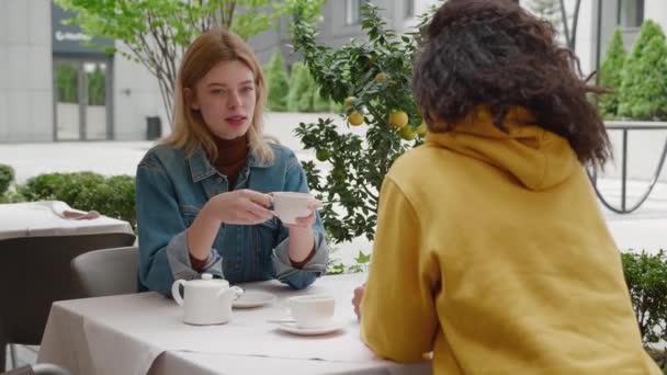 Two attractive girls sitting at the street cafe, communicate and sharing stories while, spending relax outdoor. Two diverse women friends are sitting in a cafe and talking — Stock Video