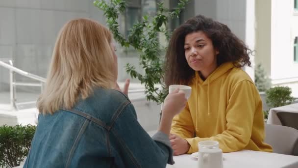 Two beautiful diverse women friends are sitting in a cafe and talking. Caucasian blonde and curly african american females friends having coffee break in cozy outdoors cafe — Stock Video