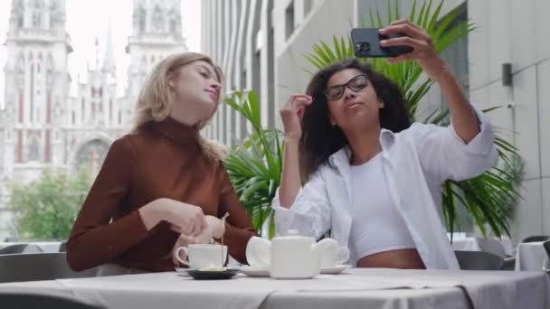 Two diverse women friends take selfie photos with mobile phone, smiling and having fun while sitting at stylish street cafe. — Stock Video