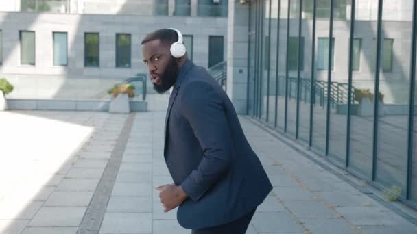 Αστείος Αφρικανός επιχειρηματίας χορεύει και τραγουδάει σε εξωτερικούς χώρους κοντά στο κέντρο γραφείου γυάλινων χώρων εργασίας φορώντας ακουστικά χαμογελώντας κρατώντας smartphone. Gadgets και έννοια ευτυχία. — Αρχείο Βίντεο