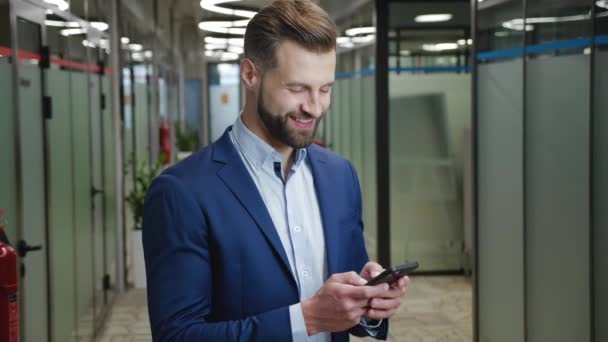 Улыбающийся бородатый бизнесмен просматривает интернет на своем смартфоне, стоя в офисном коридоре. Портрет человека в классическом костюме, печатающего сообщения на мобильном телефоне в помещении. — стоковое видео