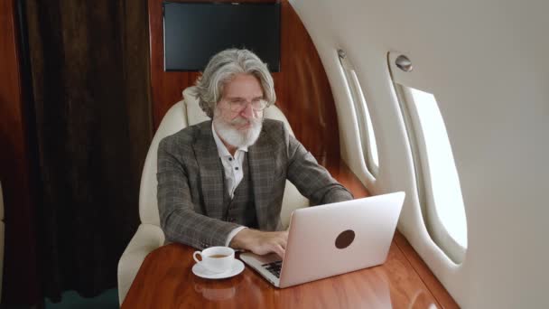 Handsome mature man millionaire working at laptop and drinking tea while traveling in airplane. Portrait of senior businessman in private jet or first business class with computer — Stock Video