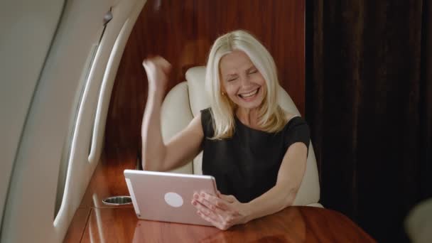 Excited mature female looks at tablet pc and rejoices in victory while flying airplane. Happy gray-haired rich senior woman celebrating success win on computer while traveling in private jet — стоковое видео
