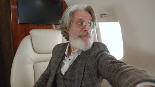 Happy mature millionaire man talking to camera holding phone recording vlog while flying on private jet. Gray-haired stylish senior man video calling in online chat using smartphone at airplane. — Stock Video