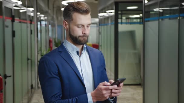 Счастливый бородатый бизнесмен наслаждается просмотром интернета на своем смартфоне, стоя в офисном коридоре. Портрет человека в классическом костюме, печатающего сообщения на мобильном телефоне в помещении. — стоковое видео