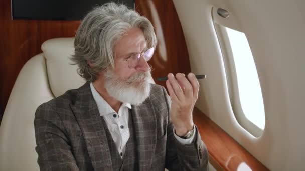 Старший чоловік мільйонер розмовляє по телефону або записує голосове повідомлення на своєму смартфоні під час польоту на приватному літаку. Багатий чоловік розмовляє по мобільному телефону під час польоту на літаку першого класу — стокове відео