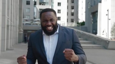 Şaşırtıcı afro-amerikan iş adamı galibiyeti kutluyor, kameraya şaşırıyor, iş bölgesinde duruyor. Takım elbiseli mutlu erkek profesyonel yönetici evet kazanan hareketi gösteriyor. Zafer konsepti