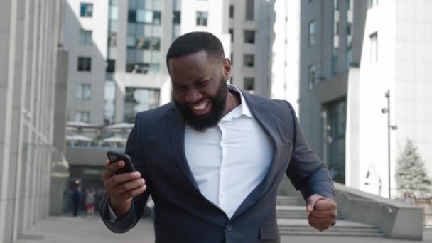 惊喜交集的非洲商人收到消息读好消息.兴奋地欣喜若狂地庆祝胜利，看着站在外面的智能手机。移动胜利概念 — 图库视频影像
