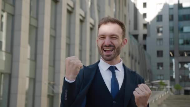 成功を祝う素晴らしいヨーロッパのビジネスマンの勝者,カメラに驚いて,ビジネス街に立って.はい勝者のジェスチャーを示す正式な訴訟で幸せな男性マネージャー。勝利のコンセプト. — ストック動画