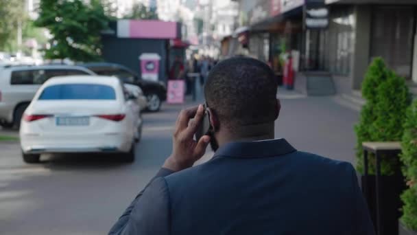 Visão traseira do homem africano bem sucedido em terno formal andando na rua da cidade falando por telefone celular. A paisagem urbana. Voltar ver empresário confiante atende o telefone fora enquanto viaja para o trabalho — Vídeo de Stock