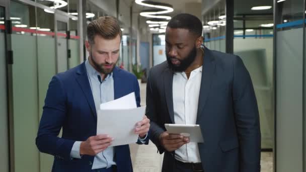 两个形式多样、快乐的生意人手握纸质文件和平板电脑，站在办公室走廊上谈论着生意。在室内谈话的男性创业者微笑 — 图库视频影像