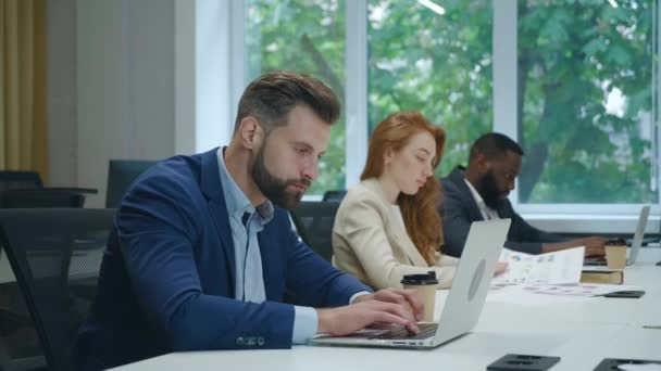 Konzentrierter Unternehmer tippt am Laptop. Junge hübsche männliche Profi mit Computer sitzt am Schreibtisch mit geschäftigen Kollegen im Hintergrund. — Stockvideo