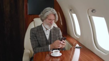 Takım elbiseli kıdemli adam özel uçakla uçarken akıllı telefon kullanıyor. Şık, olgun bir milyoner özel jetiyle uçarken cep telefonunu karıştırıyor.