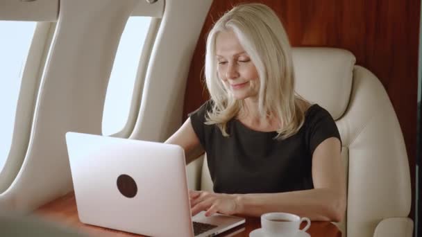 Красивая зрелая женщина-миллионер в самолете работает за компьютером ноутбука и пить кофе. Деловая женщина в частном самолете или первый бизнес-класс с компьютером. Путешествие класса люкс. — стоковое видео