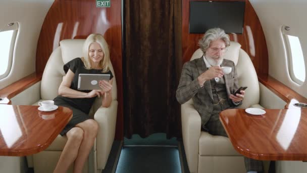 Зрелая богатая пара, использующая смартфон и планшет, путешествуя на частном реактивном самолете. Старший мужчина и женщина с помощью гаджетов, глядя в окно во время полета на авиакомпании первого класса — стоковое видео