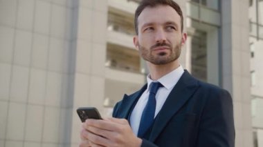 Modern Office binasının yakınındaki şehir merkezinde duran akıllı telefonu kullanan çekici iş adamının portresi. Klasik takım elbise giyiyordu. Sosyal ağ. Uygulamalar. Akıllı telefonlar.