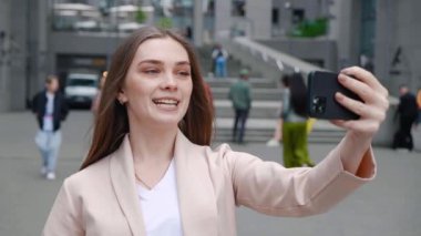 Genç ve mutlu bir iş kadını, ofis binasının dışında akıllı telefon kullanarak arkadaşlarıyla sohbet ediyor. Güzel kız öğrenci kentin arka planında beklerken selfie çekiyor.