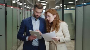 Kadın ve erkek ofis çalışanları sohbet ediyor ve tablet ve kağıt belgelerle ayakta duruyorlar. İş adamları resmi olarak ofis koridorunda dururken iş stratejilerini tartışıyor