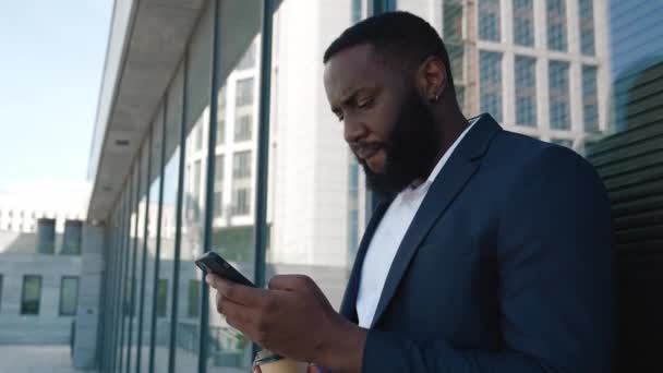 Confident bearded african american businessman browsing internet on his smartphone while standing downtown near office center. Portrait of man in classical suit reading news on mobile phone outdoors — 图库视频影像