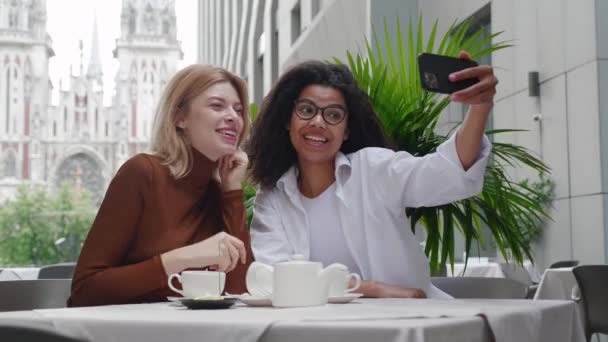 Dos jóvenes hermosas amigas que tienen chat de video selfie usando un teléfono inteligente mientras están sentadas en la cafetería al aire libre. Dos hermosas mujeres diversas hablando videollamada en el teléfono móvil en la cafetería — Vídeo de stock