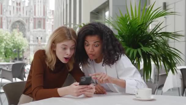 Zwei Freundinnen genießen den Erfolg mit dem Handy im Straßencafé. Überraschte Mädchen feiern den Sieg auf dem Smartphone, zeigen Wow-Emotionen, genießen den Gewinn, während sie im Café draußen sitzen — Stockvideo