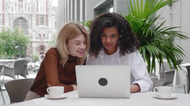 Twee vrouwelijke vrienden genieten van succes op het laptopscherm in het café. Verrast meisjes vieren overwinning op de computer, tonen wow emoties, genieten van de prijs winnen, terwijl zitten in de buitenlucht cafe — Stockvideo