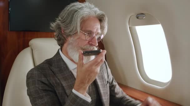 Milionario anziano che parla in vivavoce o registra un messaggio vocale sul suo smartphone mentre vola su un jet privato. Uomo ricco che parla sul cellulare mentre vola in aereo di prima classe — Video Stock