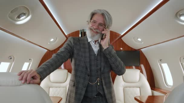Portrait d'un homme millionnaire senior élégant parlant sur un téléphone portable, debout dans un avion privé. Homme d'affaires PDG barbu souriant ayant un appel téléphonique dans un jet privé de luxe. — Video