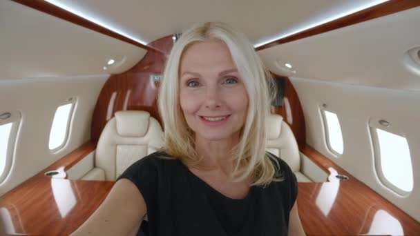POV of smiling mature businesswoman talking to camera holding phone recording vlog while standing in private jet. Gray-haired senior woman video calling in online chat using smartphone at airplane. — 图库视频影像