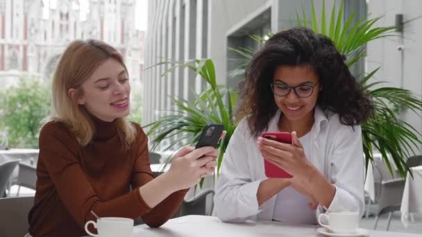 Two young beautiful women friends sitting in cafe outdoors, looking at phones and talking, laughing. Two diverse beautiful females use social media on smartphones in coffee shop, modern generation — Stock Video